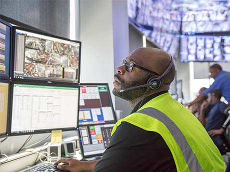 LOGSITICS TECHNOLOGY A person in a black shirt with a neon-yellow workers' vest, sits in front of a set of computer monitors and talks through a headset.