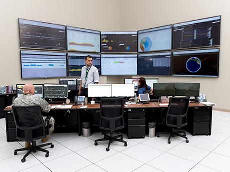 Workers sit in front of 10 large screens on the wall and multiple computer screens on a desk