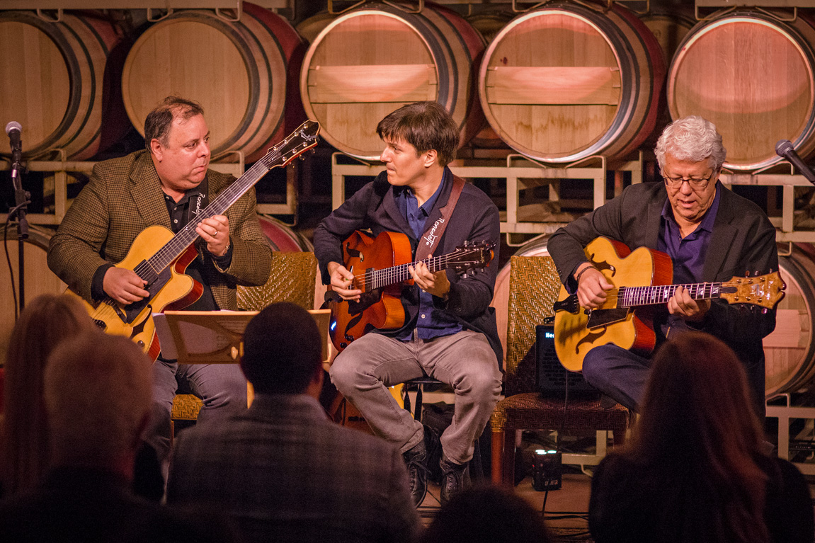 Three people on a stage play Benedetto Guitars with barrels in the background