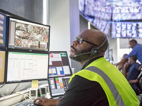 Logistics Tech A person in a black shirt with a neon-yellow workers' vest, sits in front of a set of computer monitors and talks through a headset.