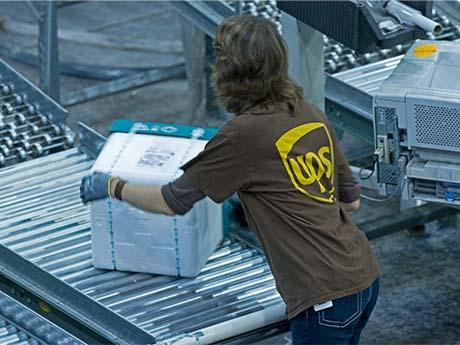 Logistics Workforce A UPS employee wearing the iconic brown and yellow uniform puts a square, white box on conveyer belt in Georgia.